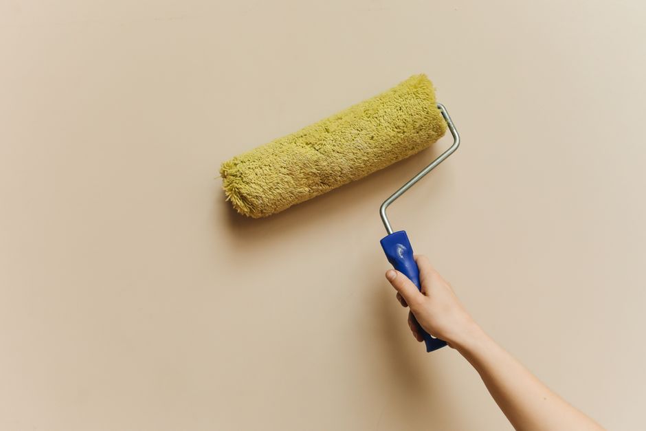 A hand using a yellow paint roller to paint a beige wall.